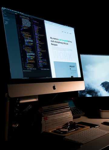 Dual monitors on a desk, one displaying code and a UX/UI designer portfolio website.