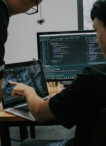 Two people reviewing computer code on a laptop and desktop monitor at a desk.